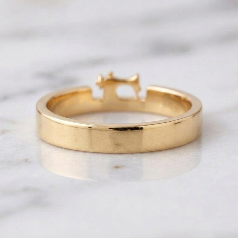 May include: A gold-toned ring featuring a small, detailed silhouette of a sewing machine integrated into the top of the band. The ring has a smooth, polished finish and a consistent, flat-profile band design that encircles the finger.
