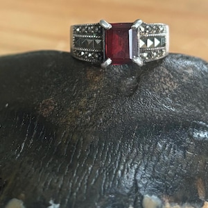 May include: A silver ring with a rectangular, deep red gemstone. The band is detailed with small, dark stones and geometric patterns. The ring is displayed on a dark, textured surface, showcasing the jewelry's design.