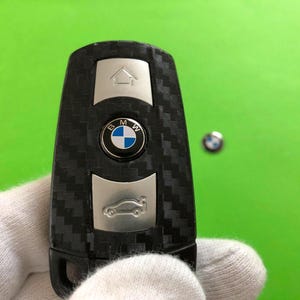 May include: A black and silver BMW car key with a carbon fiber pattern. The key has a BMW logo in the center and icons for a house and car. The key is held by a gloved hand against a green background.