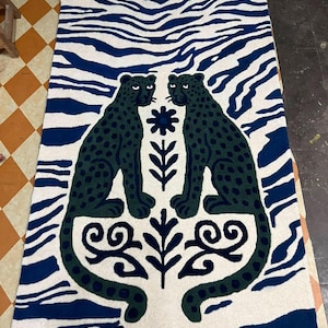 May include: A rectangular area rug featuring a cream-colored background with a blue zebra-stripe border. Two dark green spotted leopards face each other in the center, flanked by a vertical arrangement of a blue flower and leaf motifs.
