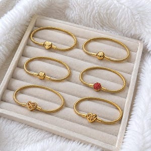Könnte beinhalten: Sechs goldfarbene Schlangenketten-Armbänder in zwei Spalten angeordnet. Jedes Armband hat einen einzigartigen Anhänger: ein glattes Herz, eine Kugelperle, eine quadratische Perle, eine rote Rose, ein Unendlichkeitsherz mit Kristallen und ein durchbrochenes Herz.