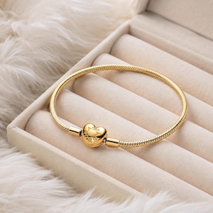 Könnte beinhalten: Ein goldenes Schlangenkettenglied-Armband mit einem herzförmigen Verschluss. Der Verschluss ist mit dem Wort PANDORA graviert. Das Armband hat ein glattes, flexibles und abgerundetes Glieddesign, das im geschlossenen Zustand einen Kreis bildet.