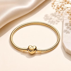 May include: A gold-toned snake chain bracelet featuring a central heart-shaped charm. The charm is embossed with a star and circular design. The bracelet has a smooth, polished finish and a flexible, rounded link structure, creating a continuous circular shape.