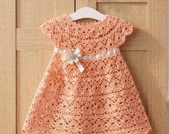 May include: A peach-colored crochet dress with a scalloped hem and cap sleeves hangs on a white hanger. A white ribbon with a bow and a decorative embellishment accents the waist. The dress is displayed against a light wood background.