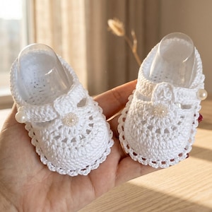 May include: A pair of white crochet baby shoes. Each shoe has an openwork design, a strap with a pearl button, and a pearl accent. The shoes are designed with a scalloped edge, perfect for a newborn.