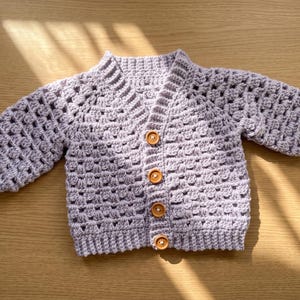 Könnte beinhalten: Ein helllila gehäkelter Baby-Cardigan mit V-Ausschnitt und langen Ärmeln. Der Pullover hat eine Knopfleiste mit vier Holzknöpfen. Das Häkelmuster ist offen und luftig. Der Cardigan liegt auf einer Holzoberfläche.