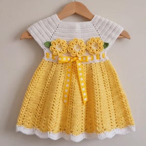 May include: A crochet dress featuring a white bodice and a yellow skirt with a textured pattern. The waist is accented with three yellow crochet flowers, green leaves, and a yellow ribbon with white polka dots. The hem is finished with white trim.
