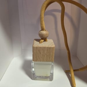 Pode incluir: Um pequeno frasco quadrado de vidro transparente preenchido com um líquido pálido, rematado com uma tampa de madeira clara. Uma conta de madeira redonda prende um cordão dourado que passa pela tampa, permitindo pendurar o frasco como ambientador.
