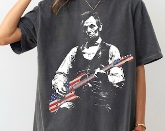 May include: Dark gray t-shirt featuring a black and white image of Abraham Lincoln playing an electric guitar with an American flag design. The shirt has short sleeves and a relaxed fit.