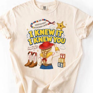 May include: A cream-coloured t-shirt featuring a graphic print of a person with a blonde braid wearing a red cowboy hat, white shirt, and blue jeans. The text reads I KNEW IT I KNEW YOU, YOU'VE GOT A FRIEND IN ME, and IT'S A LOVE STORY BABY JUST SAY YES.