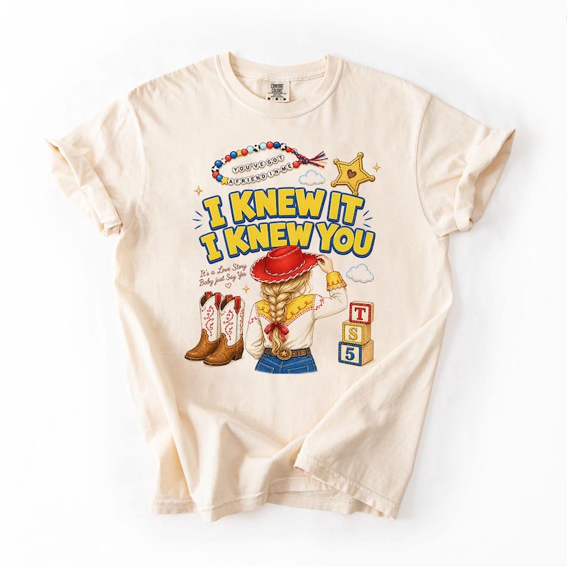 May include: A cream-coloured t-shirt featuring a graphic print of a person with a blonde braid wearing a red cowboy hat, white shirt, and blue jeans. The text reads I KNEW IT I KNEW YOU, YOU'VE GOT A FRIEND IN ME, and IT'S A LOVE STORY BABY JUST SAY YES.
