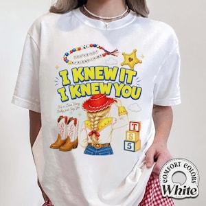 May include: A white t-shirt featuring a graphic print of a person with blonde hair in a braid wearing a red cowboy hat, brown cowboy boots, and a yellow shirt. The text reads I KNEW IT I KNEW YOU, YOU'VE GOT A FRIEND IN ME, and It's a Love Story Baby just Say Yes.
