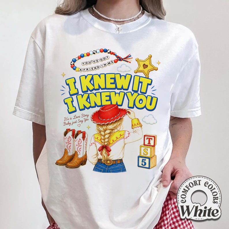 May include: A white t-shirt featuring a graphic print of a person with blonde hair in a braid wearing a red cowboy hat, brown cowboy boots, and a yellow shirt. The text reads I KNEW IT I KNEW YOU, YOU'VE GOT A FRIEND IN ME, and It's a Love Story Baby just Say Yes.