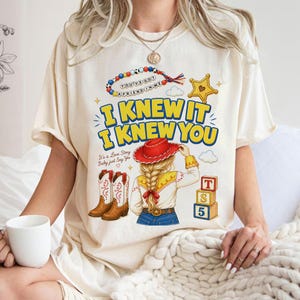 May include: A cream-coloured short-sleeve t-shirt featuring a graphic print with the text I KNEW IT I KNEW YOU, a beaded necklace illustration, a cowboy hat, boots, and blocks. The design includes a person with blonde hair in a braid.