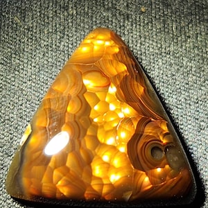 May include: A triangular-shaped polished gemstone featuring translucent amber and golden-brown tones with intricate, concentric banding patterns and a glowing, honey-like internal structure that catches the light across its smooth, rounded surface.