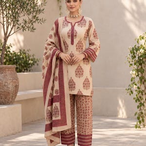 May include: A beige three-piece outfit consisting of a long-sleeved tunic, wide-leg trousers, and a matching shawl. The garments feature a repeating dark red botanical block print pattern, with solid dark red bands at the cuffs and hem.
