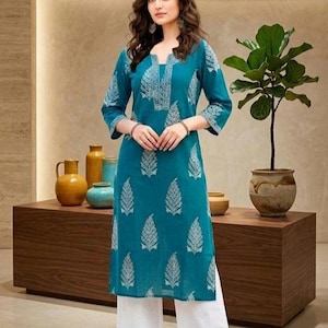 May include: A teal tunic with white leaf patterns and three-quarter sleeves, worn with white palazzo trousers. The outfit is completed with brown flats. A wooden table holds decorative vases and a potted plant.