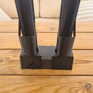 Puede incluir: Un soporte negro impreso en 3D que presenta dos tubos cil&iacute;ndricos verticales unidos a una base rectangular. La base incluye dos secciones laterales con un patr&oacute;n de panal perforado y un bloque central s&oacute;lido que conecta los dos soportes.