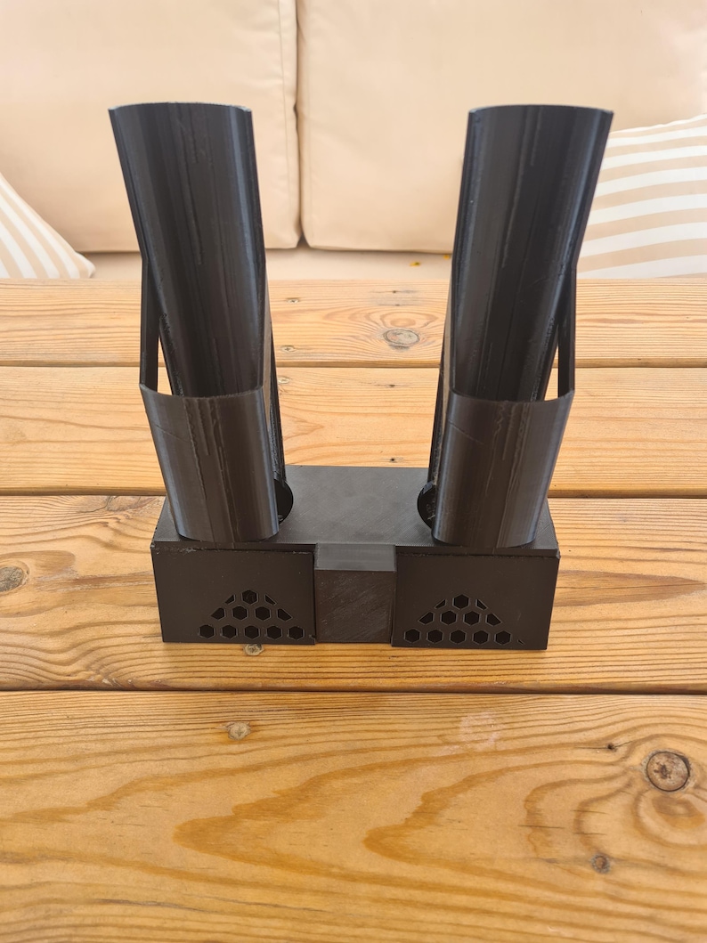 Puede incluir: Un soporte negro impreso en 3D que presenta dos tubos cil&iacute;ndricos verticales unidos a una base rectangular. La base incluye dos secciones laterales con un patr&oacute;n de panal perforado y un bloque central s&oacute;lido que conecta los dos soportes.