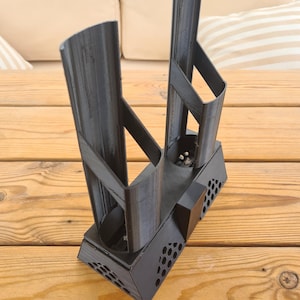 Puede incluir: Un soporte negro impreso en 3D con dos soportes verticales semicil&iacute;ndricos unidos a una base rectangular. La base tiene un panel lateral texturizado con patr&oacute;n de panal y un bloque central, con l&iacute;neas de capa visibles en toda la estructura.