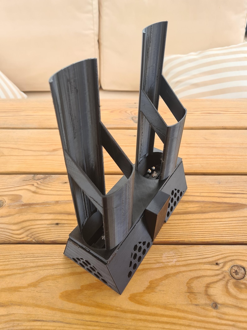 Puede incluir: Un soporte negro impreso en 3D con dos soportes verticales semicil&iacute;ndricos unidos a una base rectangular. La base tiene un panel lateral texturizado con patr&oacute;n de panal y un bloque central, con l&iacute;neas de capa visibles en toda la estructura.