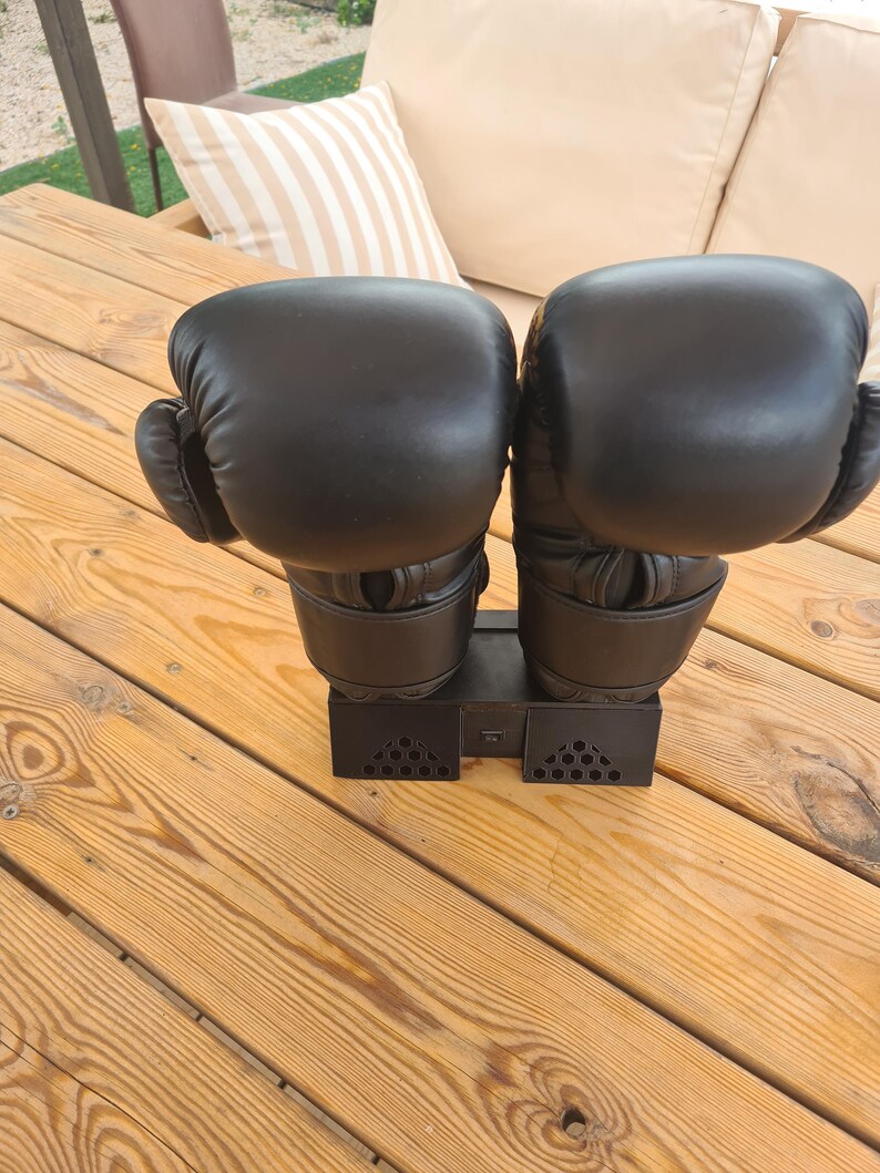 Puede incluir: Un par de guantes de boxeo negros montados sobre un soporte de pl&aacute;stico negro. Los guantes est&aacute;n colocados uno al lado del otro, con un acabado liso y mate y costuras visibles en la zona de la mu&ntilde;eca. El soporte tiene una base rectangular.