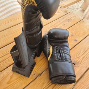 Puede incluir: Un par de guantes de boxeo negros con detalles dorados en la mu&ntilde;eca y el pulgar. Un guante est&aacute; colocado verticalmente sobre un soporte de pl&aacute;stico negro, mientras que el otro guante yace plano de lado. Los guantes tienen un acabado liso.