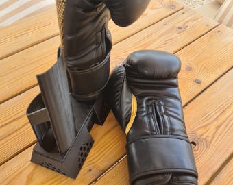 May include: A pair of black boxing gloves with gold accents on the wrist and thumb area. One glove is positioned upright on a black plastic stand, while the other glove lies flat on its side. The gloves feature a smooth finish and stitched details.