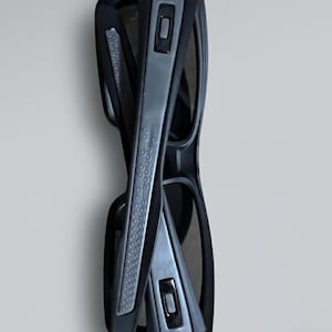 May include: Folded black sunglasses with gray accents. The sunglasses have a rectangular shape and a textured grip on the arms. The lenses are dark, and the frame is a matte black.