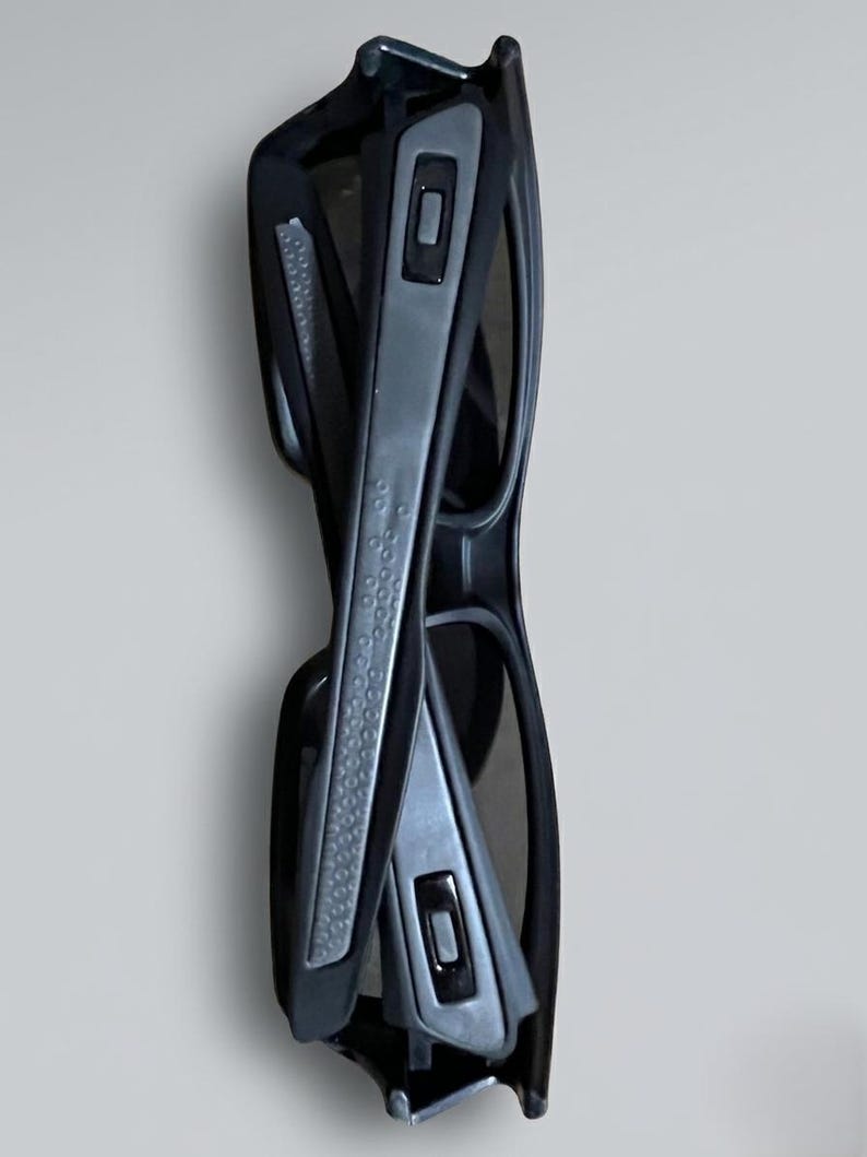 May include: Folded black sunglasses with gray accents. The sunglasses have a rectangular shape and a textured grip on the arms. The lenses are dark, and the frame is a matte black.