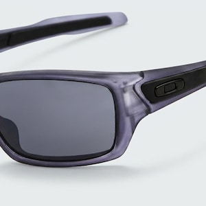 May include: Gray sunglasses with dark lenses. The frames are a translucent gray with black accents on the temples and hinges. The sunglasses have a sporty, rectangular shape and are designed for outdoor activities.