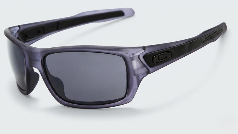 May include: Gray sunglasses with dark lenses. The frames are a translucent gray with black accents on the temples and hinges. The sunglasses have a sporty, rectangular shape and are designed for outdoor activities.