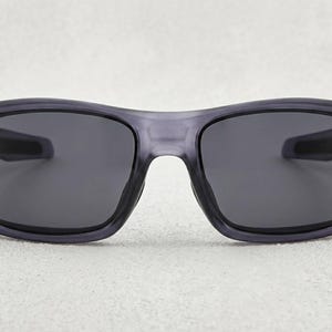 May include: Gray sunglasses with dark lenses. The frames are a translucent gray color, and the lenses are a solid dark gray. The sunglasses have a rectangular shape with a slightly curved top and are designed for eye protection.