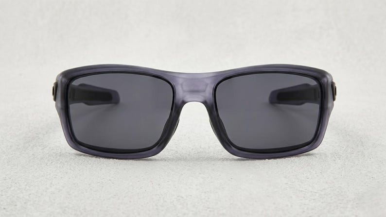 May include: Gray sunglasses with dark lenses. The frames are a translucent gray color, and the lenses are a solid dark gray. The sunglasses have a rectangular shape with a slightly curved top and are designed for eye protection.