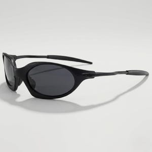 Puede incluir: Gafas de sol negras de forma ovalada con lentes oscuras. Las gafas tienen un dise&ntilde;o elegante y moderno con un acabado mate. Las patillas son finas y se curvan ligeramente, y las lentes son de color gris oscuro.