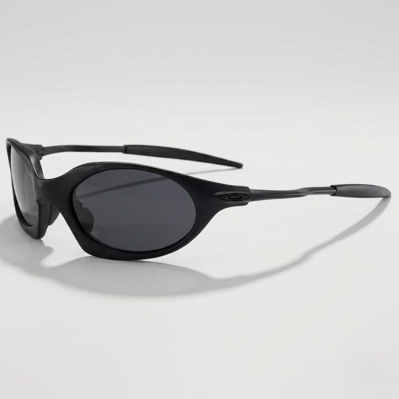 Puede incluir: Gafas de sol negras de forma ovalada con lentes oscuras. Las gafas tienen un dise&ntilde;o elegante y moderno con un acabado mate. Las patillas son finas y se curvan ligeramente, y las lentes son de color gris oscuro.
