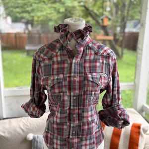 May include: A long-sleeve button-up shirt featuring a red, white, and gray plaid pattern. The shirt has a ruffled collar, two chest pockets with snap closures, and a front placket with silver-toned snap buttons. The sleeves are rolled up at the cuffs.
