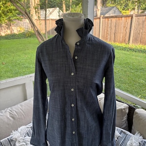 May include: A dark blue long-sleeve button-down shirt with a textured fabric finish. The shirt features a high, ruffled collar and a front placket with white buttons. The sleeves are long with buttoned cuffs, and the overall design is simple.