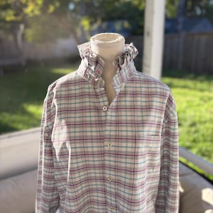 May include: A long-sleeved button-down shirt featuring a plaid pattern in shades of cream, magenta, and teal. The shirt includes a distinctive ruffled collar, a front button placket with light-colored buttons, and a single chest pocket on the left side.
