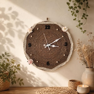 May include: A round wall clock featuring a dark brown textured jute or twine surface. The clock face includes black numerals at 12, 3, 6, and 9, white hands, and small white beads as markers. A white lace trim edges the clock, accented with three pink flowers.