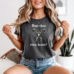 May include: A dark gray short-sleeve t-shirt featuring a graphic of a knight in armor lifting a barbell. The text "Dost thou even heave?" is printed in white gothic-style lettering above and below the knight illustration on the chest.