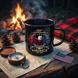 Puede incluir: Una taza de esmalte negro con un dise&ntilde;o patri&oacute;tico que presenta un &aacute;guila calva, el texto "250th Anniversary AMERICA 1776-2026" y la bandera estadounidense. La taza est&aacute; sobre un mapa con una br&uacute;jula, pi&ntilde;as y una fogata en el fondo.