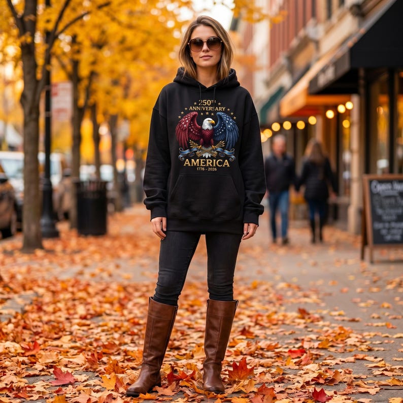 Puede incluir: Sudadera con capucha negra con un dise&ntilde;o de &aacute;guila con alas rojas y azules, el texto "250th Anniversary AMERICA 1776-2026". La persona lleva vaqueros negros y botas marrones, de pie en una calle con hojas de oto&ntilde;o.
