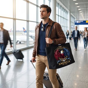 Puede incluir: Bolso tote negro con un dise&ntilde;o patri&oacute;tico de un &aacute;guila roja, blanca y azul, estrellas y el texto "250th Anniversary AMERICA 1776-2026". La persona lleva el bolso en un aeropuerto.