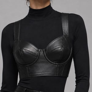 May include: A black leather bustier top with wide shoulder straps and structured cups, worn over a black long-sleeved mock neck shirt. The bustier features visible seam detailing across the front and a cropped hemline that sits above the waist.