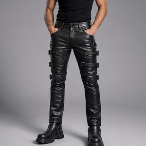 May include: A black sleeveless ribbed tank top paired with black leather pants featuring multiple horizontal straps, metal buckles, and D-ring hardware along the thighs and shins, finished with black leather boots with thick lug soles.