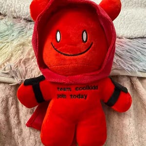 May include: A plush red doll with a hood and small horns. The doll has a smiling face with white eyes and a black mouth. The text "team coolkidd join today" is embroidered on the doll's body. The doll has black armbands and a small cape.
