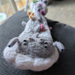 Può includere: Una creatura fatta a mano in feltro grigio con più occhi e piccole decorazioni colorate. La creatura ha una pancia bianca e piccole antenne grigie. Gli occhi sono di vari colori, tra cui blu, verde e oro. La creatura è appoggiata su una superficie grigio scuro.