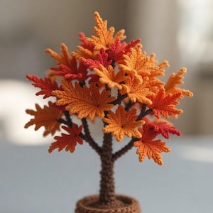 May include: A crocheted maple tree sculpture featuring a brown textured trunk and base with vibrant orange and red autumn leaves. The small tree is crafted with detailed yarn stitches, showcasing a dense canopy of pointed, serrated foliage.