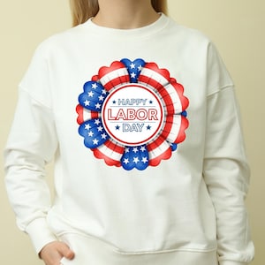 Könnte beinhalten: Weißes Sweatshirt mit einem kreisförmigen Design mit dem Text "HAPPY LABOR DAY" in einem rot-weißen Kreis, umgeben von einem blau-weiß-roten Rosettendesign. Das Design erinnert an die amerikanische Flagge.